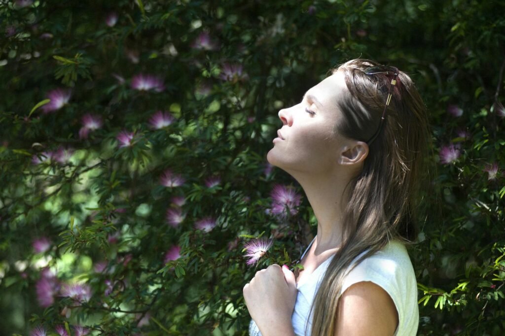 Why Self-Compassion for Parents Is the Key to Better Parenting woman closing her eyes against sun light standing near purple petaled flower plant